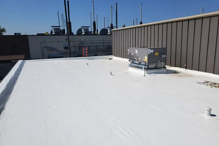 White flat commercial roof with HVAC unit, various pipes, and antennas in the background.
