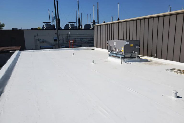 White flat roof with HVAC unit and various pipes under a blue sky.