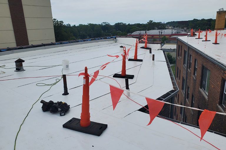 Workers on a flat white roof, with orange safety cones, flags, and wires.