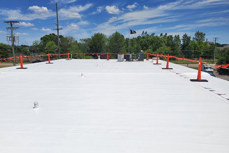 White commercial roof with orange safety cones, clear blue sky, and trees in background.