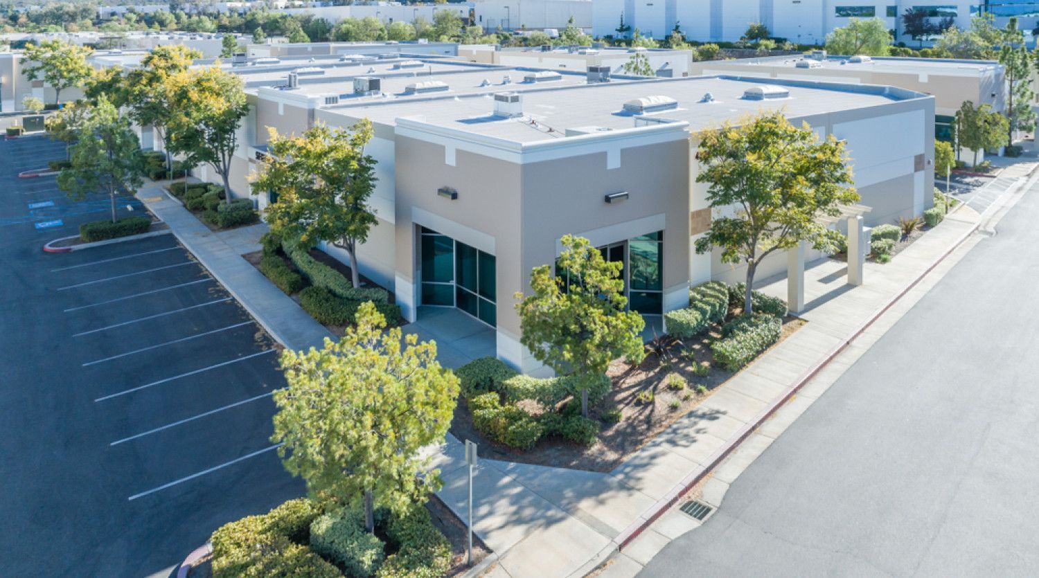 Industrial building with tan and gray facade, trees, and parking lot.