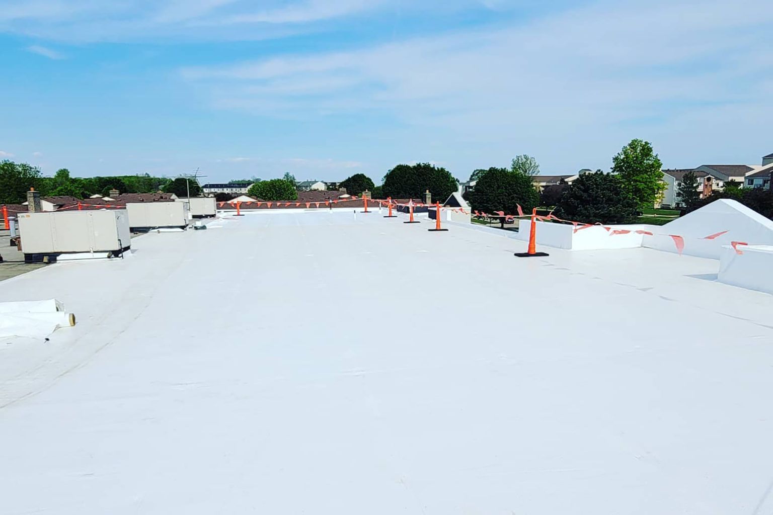 White commercial roof being worked on with trucks and cones in the background. Blue sky.