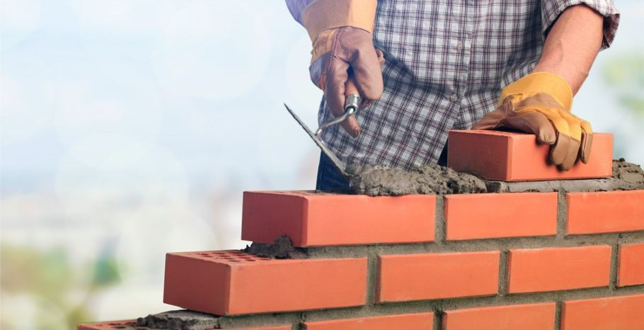 Worker laying red bricks on a wall with mortar, wearing gloves and a checkered shirt