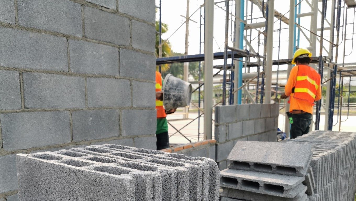 Construction workers in orange vests building a block wall on scaffolding at a site.