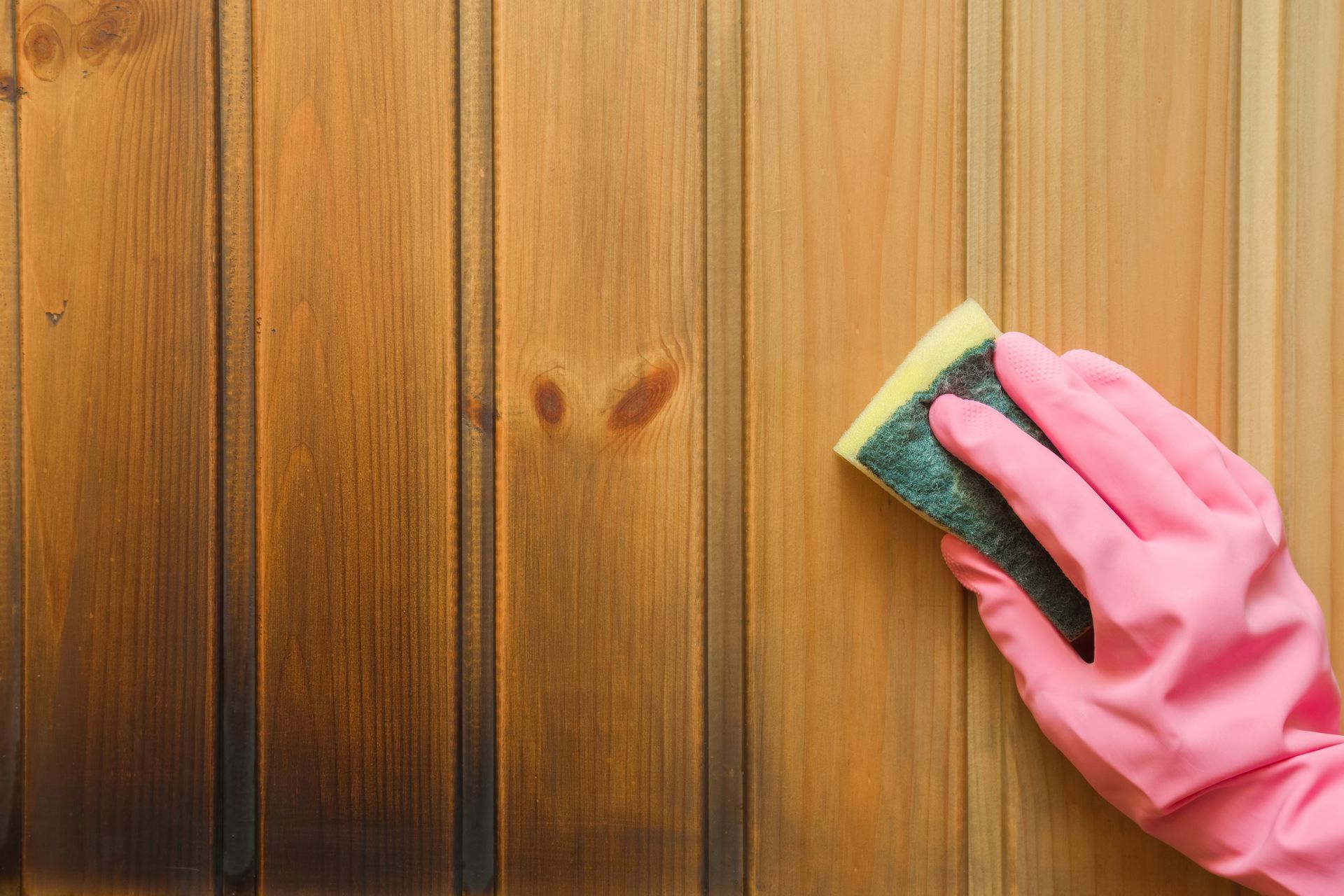 Pink-gloved hand scrubbing a wooden wall with a green sponge