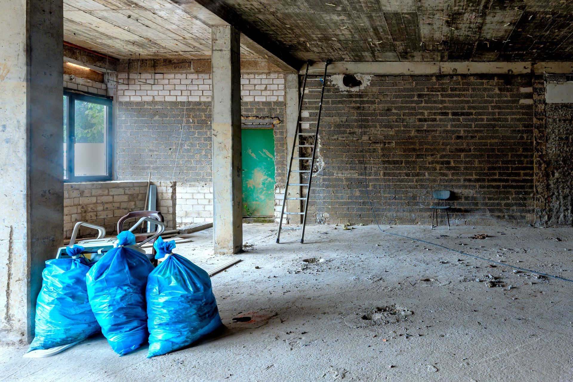 Abandoned unfinished room with blue trash bags, concrete columns, and a green door.