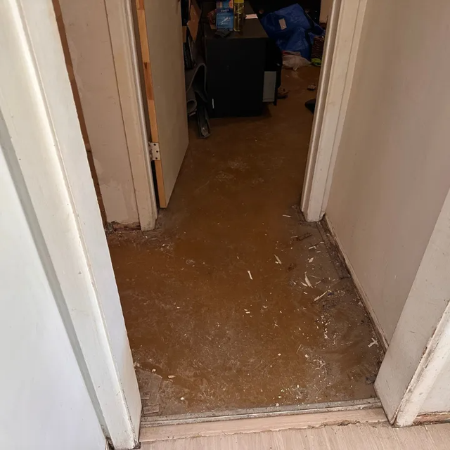 Narrow hallway with white walls and a brown, debris-covered floor leading to a cluttered room