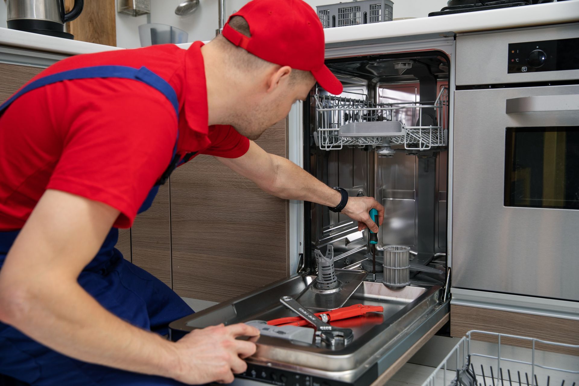 Technician in red cap and blue overalls repairing a dishwasher with tools in a kitchen.
