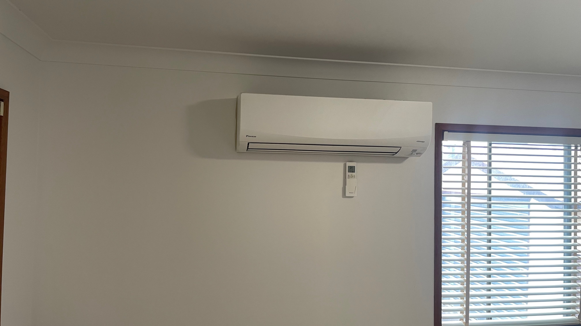 White wall with air conditioner and remote; window with blinds.