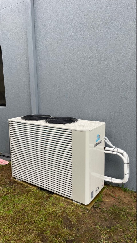 White rectangular pool heat pump next to a gray building. Two black fans sit on top.