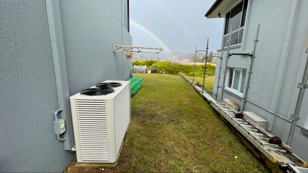 Air conditioning unit next to a gray building. Rainbow in the background over green grass.