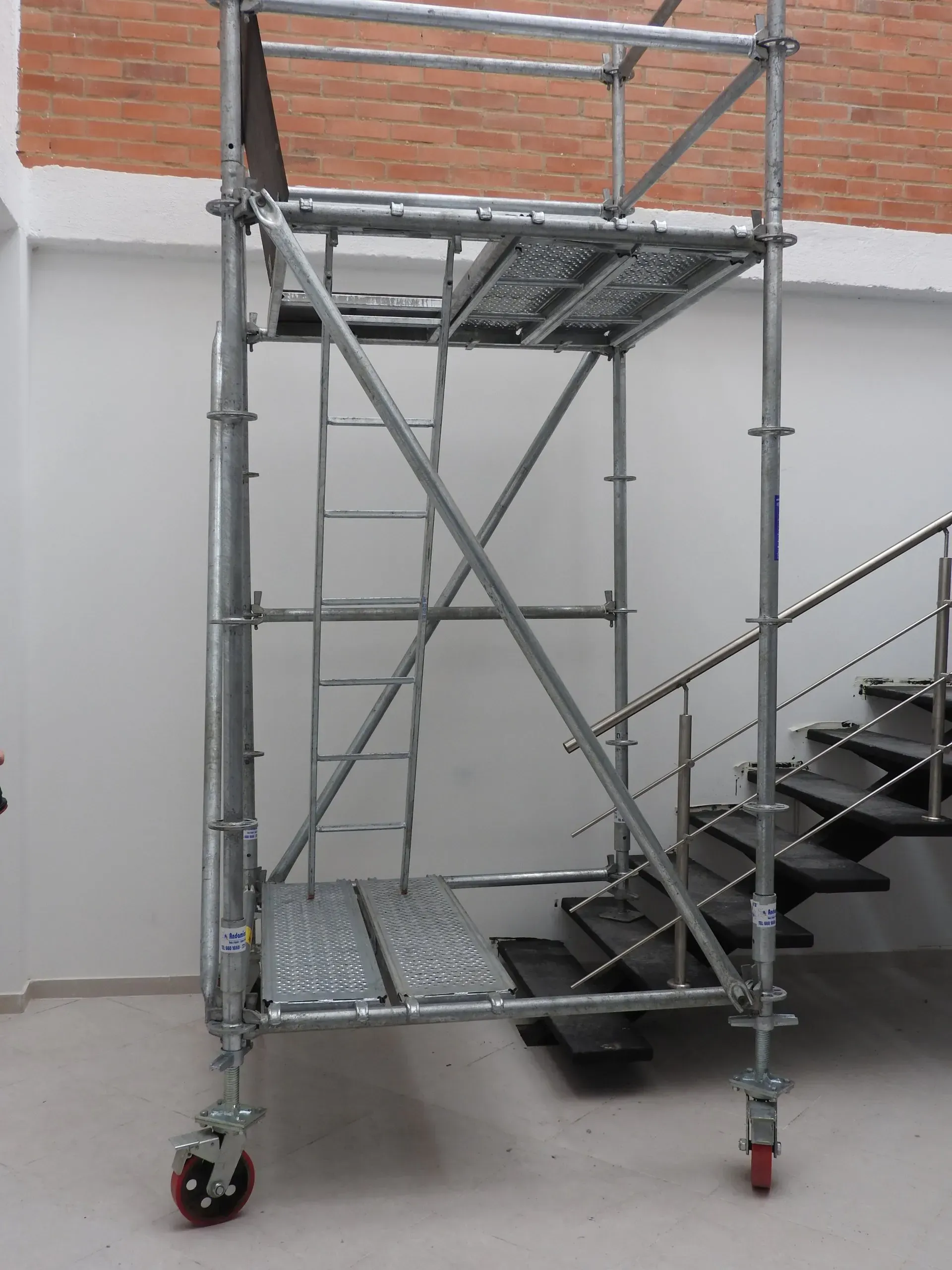 Torre móvil de andamio con plataforma metálica y peldaños junto a una pared de ladrillo en interiores