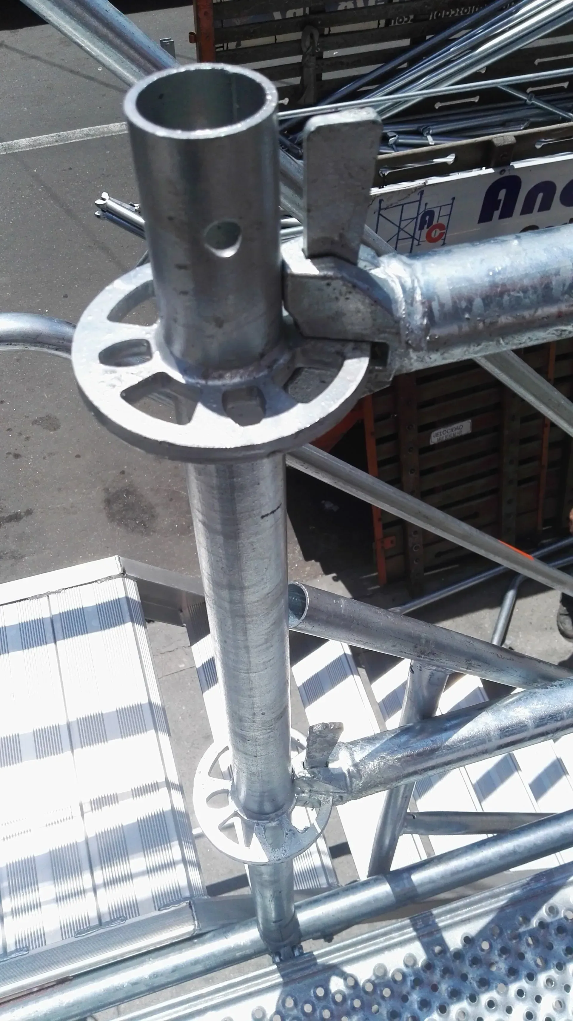 Unión metálica de andamio con una placa de sujeción blanca y un poste vertical plateado en una obra en construcción.