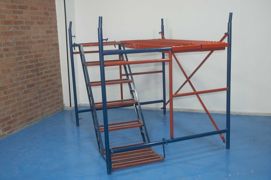 Andamio azul y naranja con una escalera en una habitación vacía, junto a una pared de ladrillo