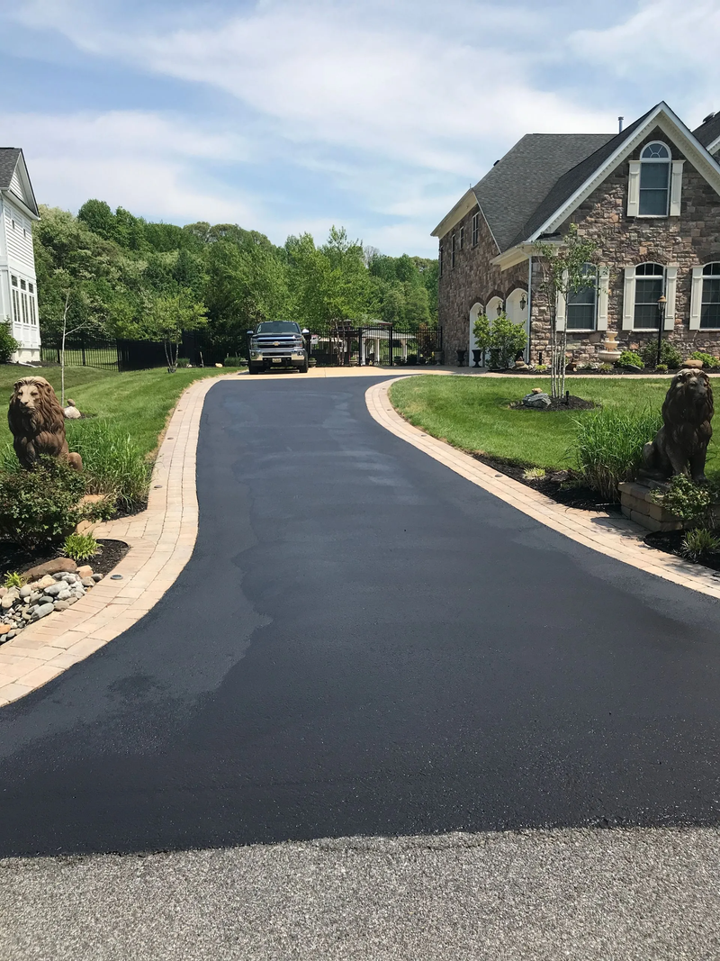 Fresh black asphalt driveway curving toward a stone house with landscaped greenery under a blue sky