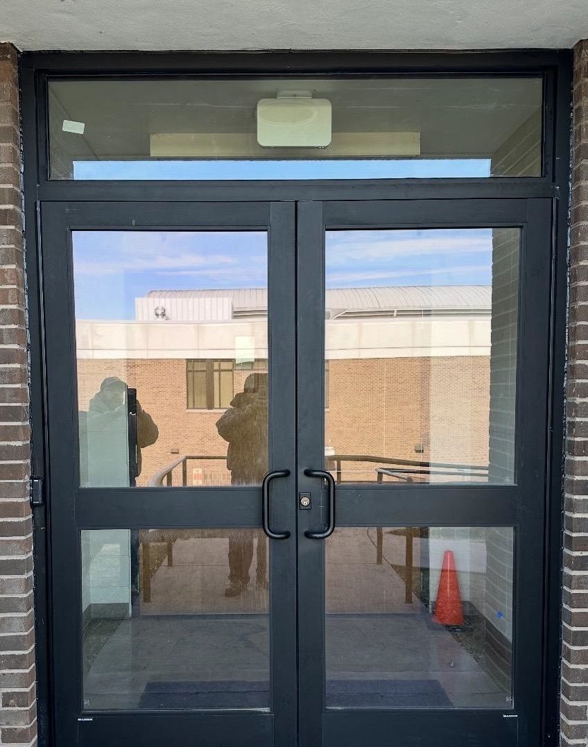 Double glass doors with black frames in a brick entryway; reflections of buildings and person visible.