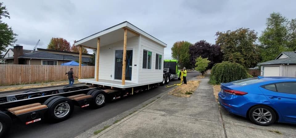 Tiny house on a flatbed trailer being towed along a driveway, with a blue car nearby.