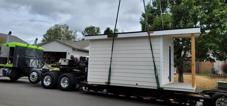 Flatbed truck hauling a white portable building with an open doorway along a street