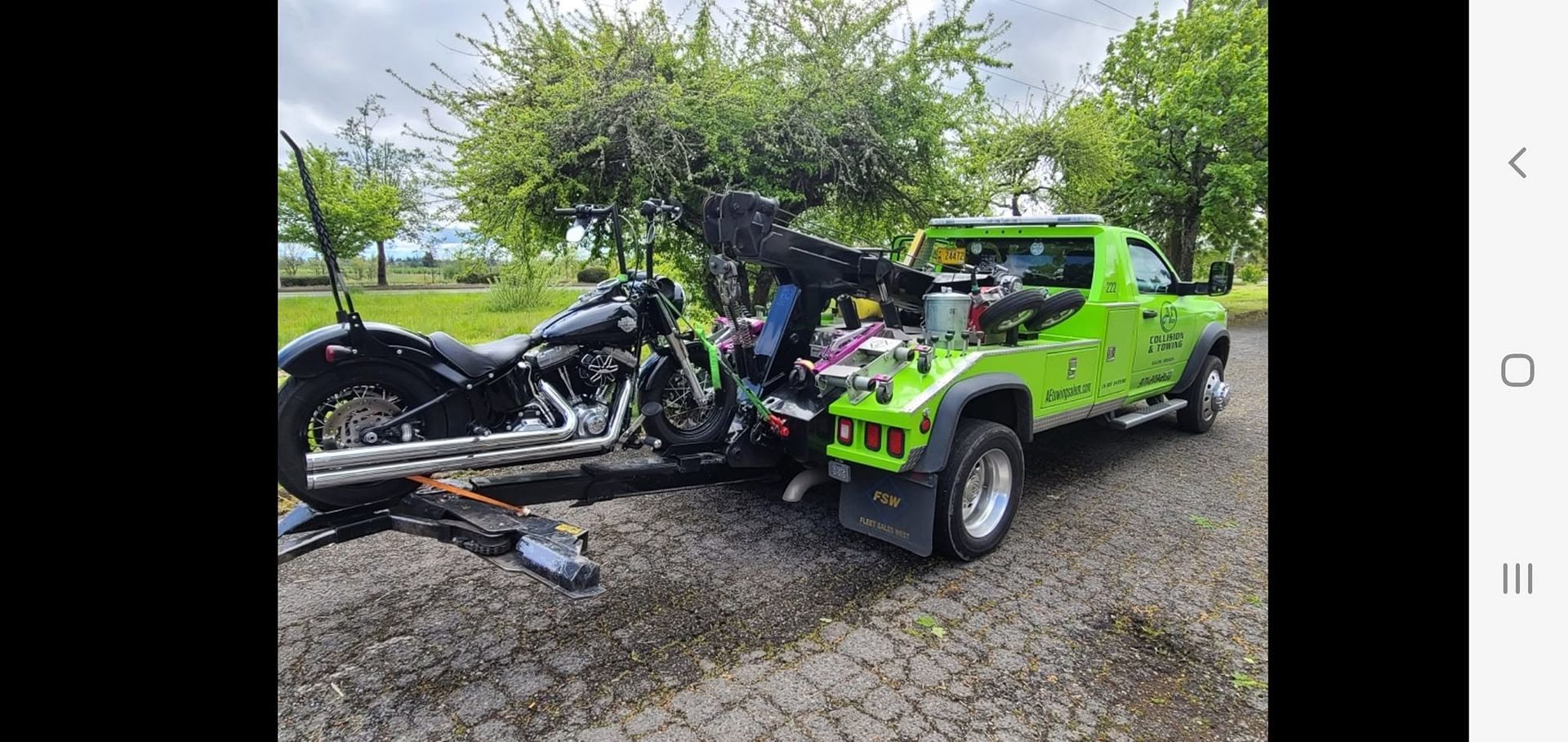 Bright green tow truck carrying a mangled black car on a wet gravel roadside under cloudy skies