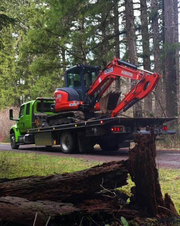 Green flatbed truck hauling a red excavator beside a wooded roadside area