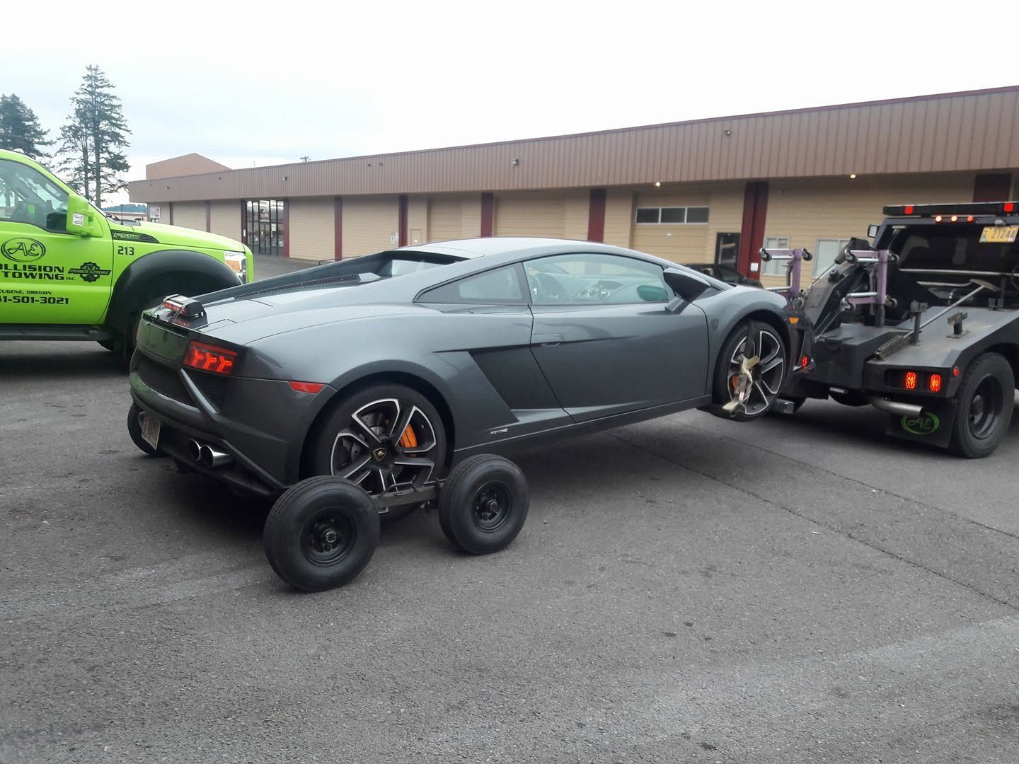 Gray sports car on a trailer with racing wheels in a parking lot, next to a green car and a tow truck
