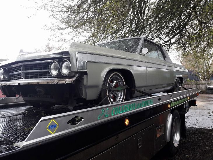 Classic silver car on a tow truck flatbed outdoors under trees.