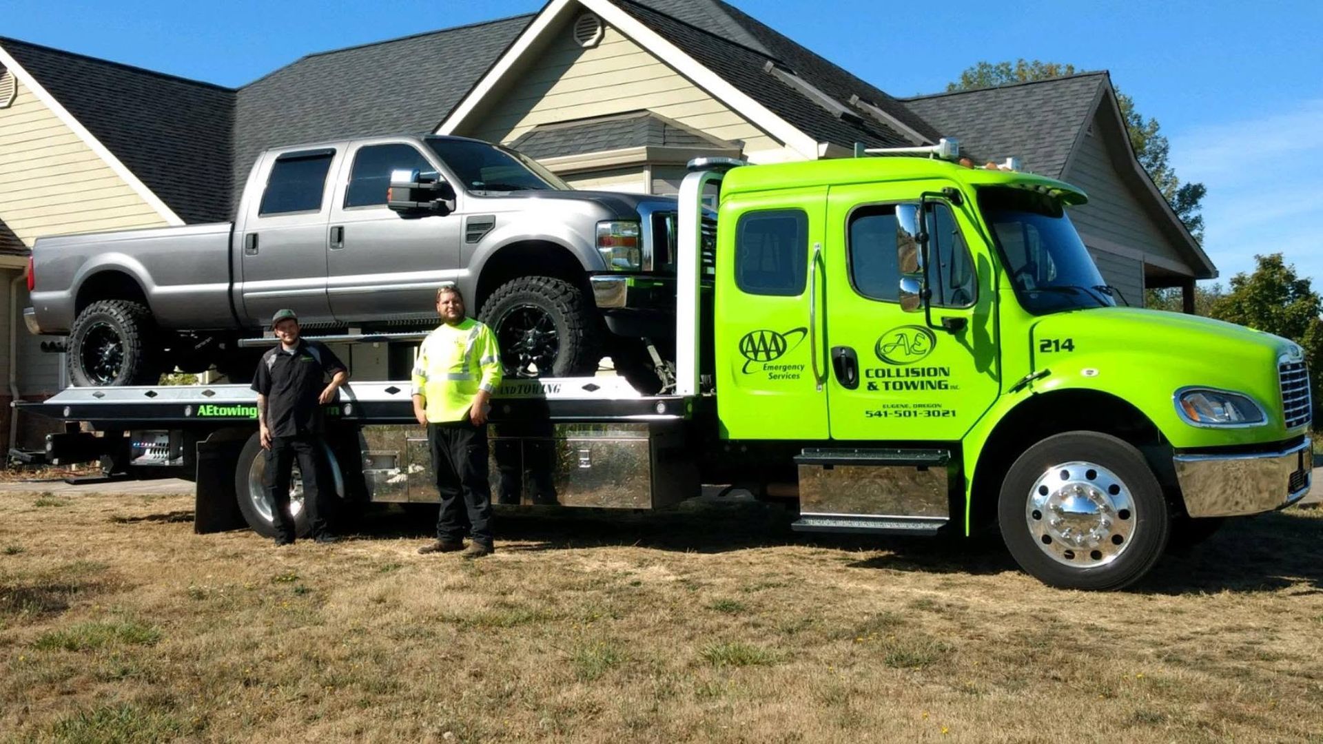 Tow truck loading a silver pickup outside a suburban house, with two workers in safety vests.