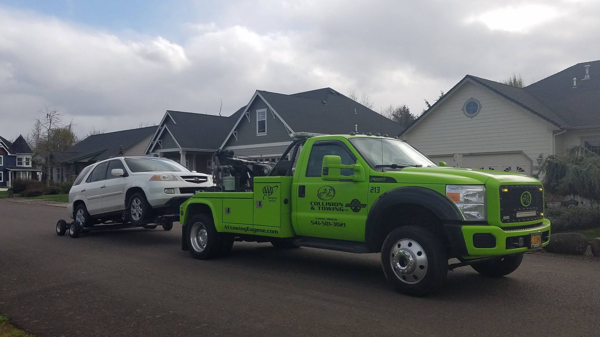 Bright green tow truck carrying a white SUV on a residential street under cloudy skies