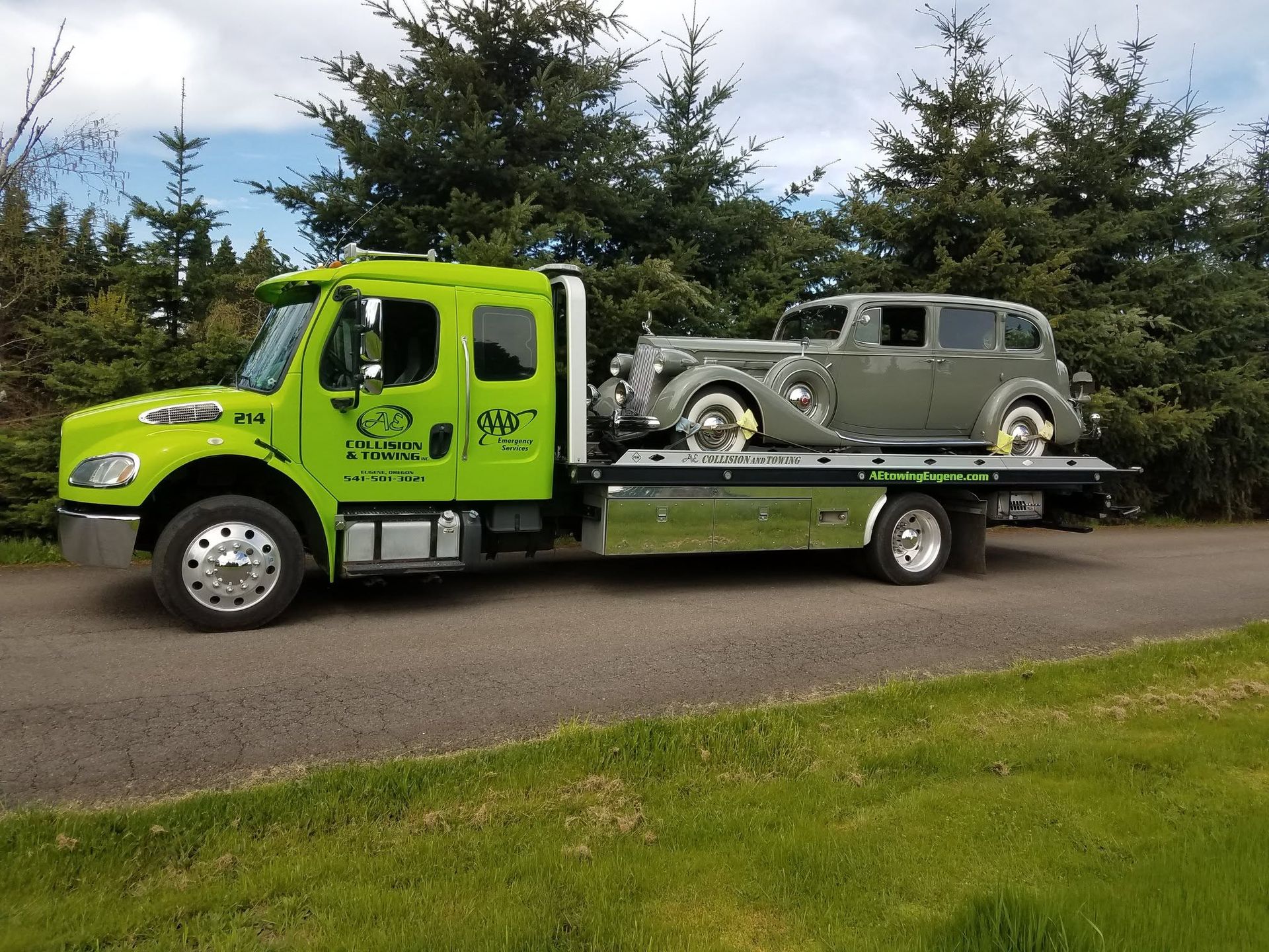 Bright green tow truck carrying a gray vintage car on a flatbed by a roadside.