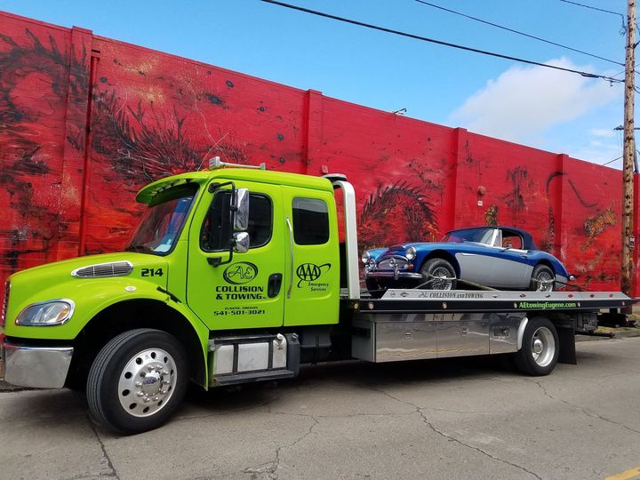 Bright green tow truck hauling a blue convertible on a flatbed beside a red wall