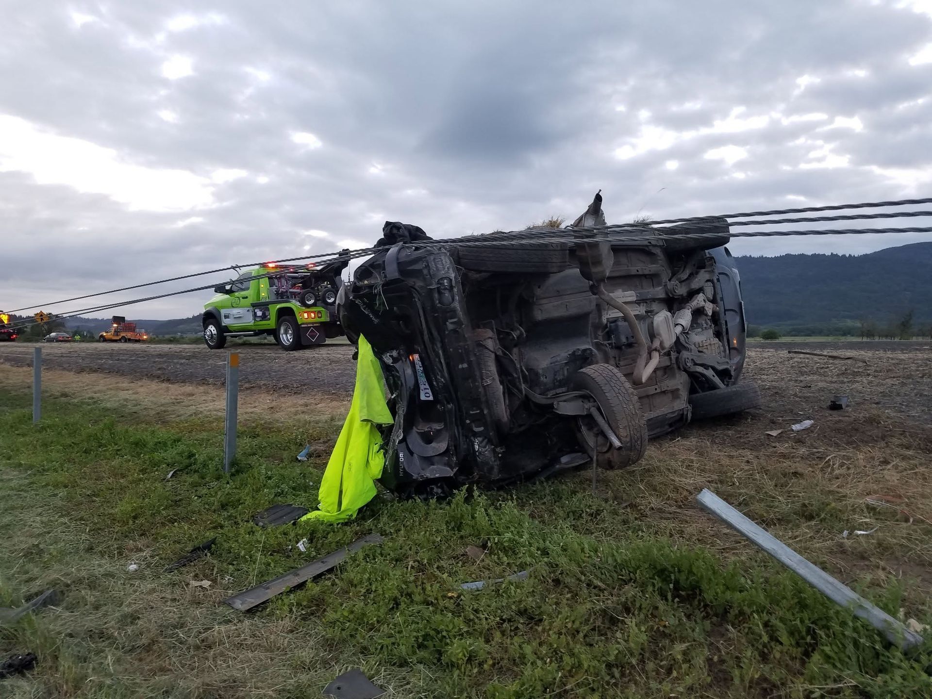 Overturned black vehicle in a roadside crash, with tow truck and emergency crew nearby under cloudy sky