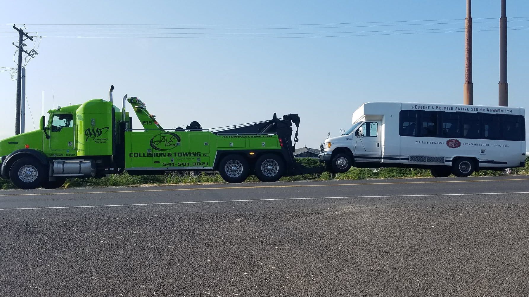 Green tow truck hauling a white shuttle bus beside a gravel roadside under a clear blue sky
