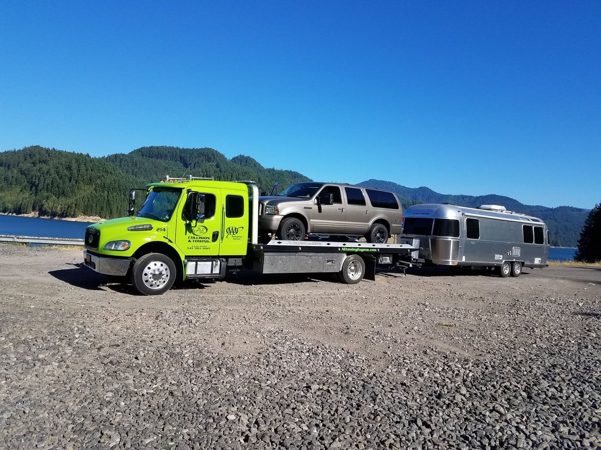 Bright green tow truck hauling a silver pickup and trailer across a gravel lot with mountains in the background