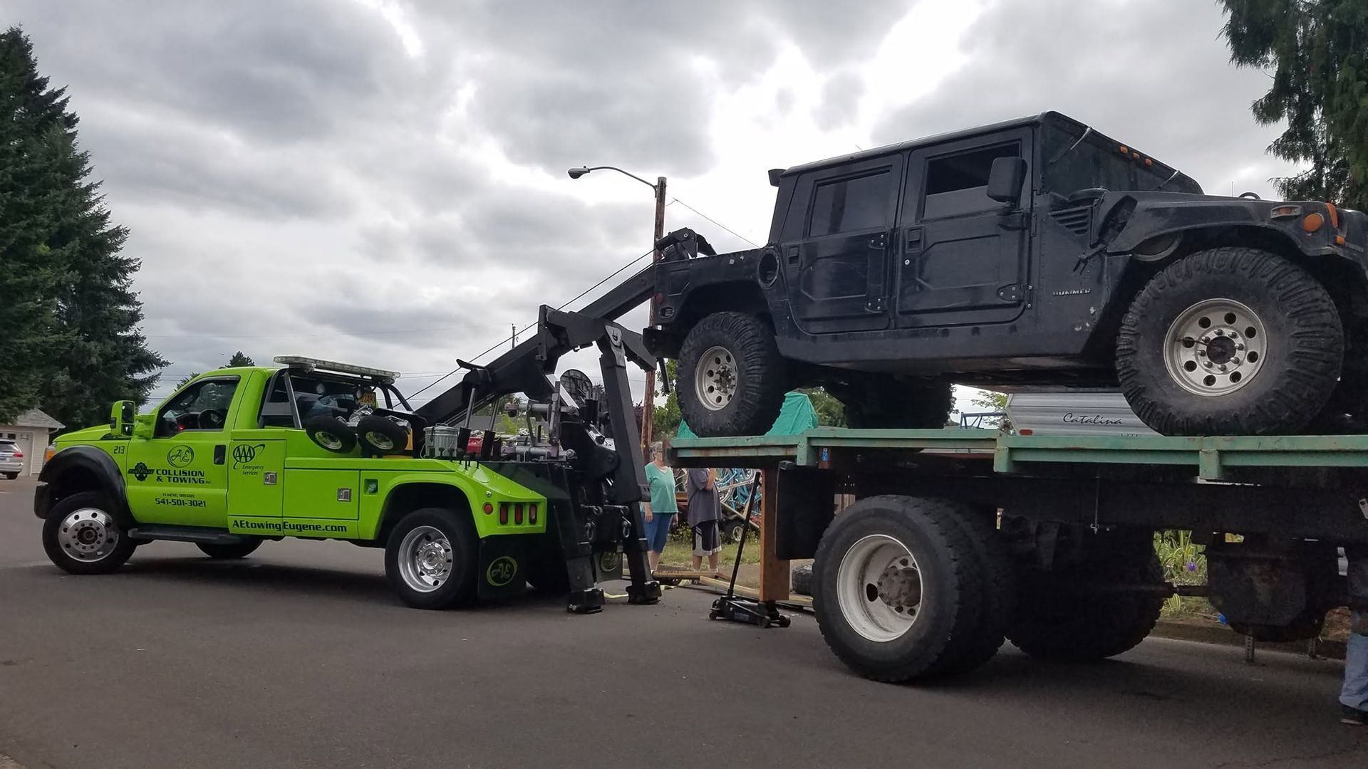 Tow truck lifting a black off-road Jeep beside a bright green pickup under a cloudy sky