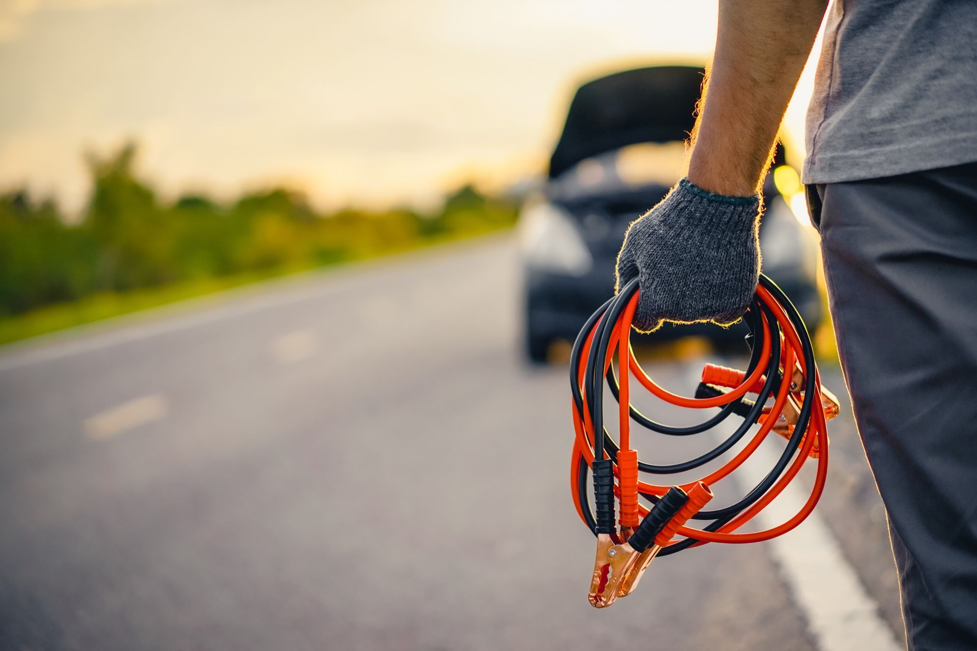 Gloved person holding jumper cables beside a broken-down car on a roadside