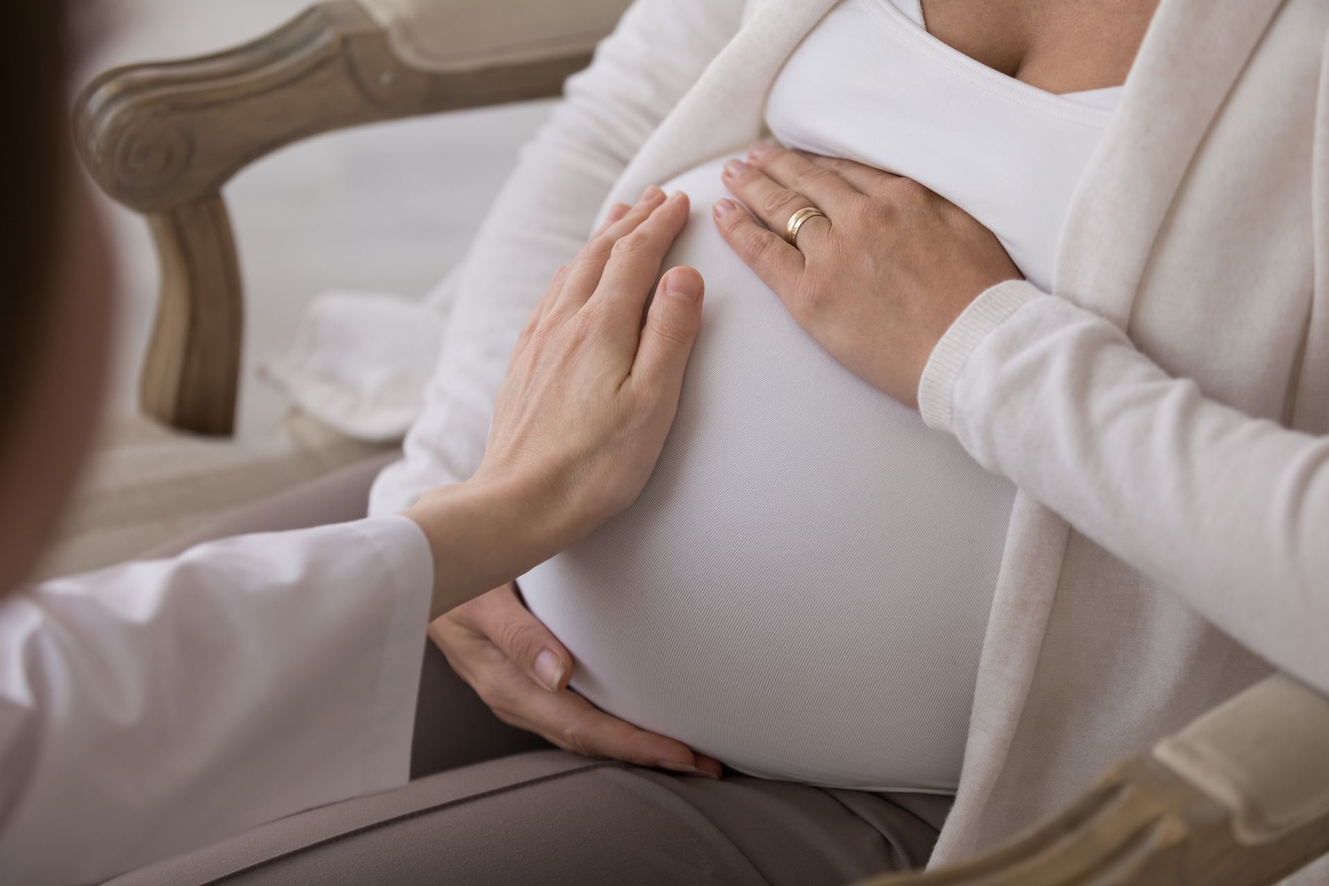Pregnant woman seated indoors while a healthcare professional places a hand on her abdomen.