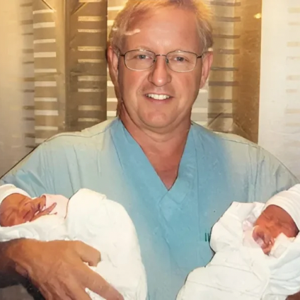 Doctor in scrubs holds newborn twins in a hospital setting.