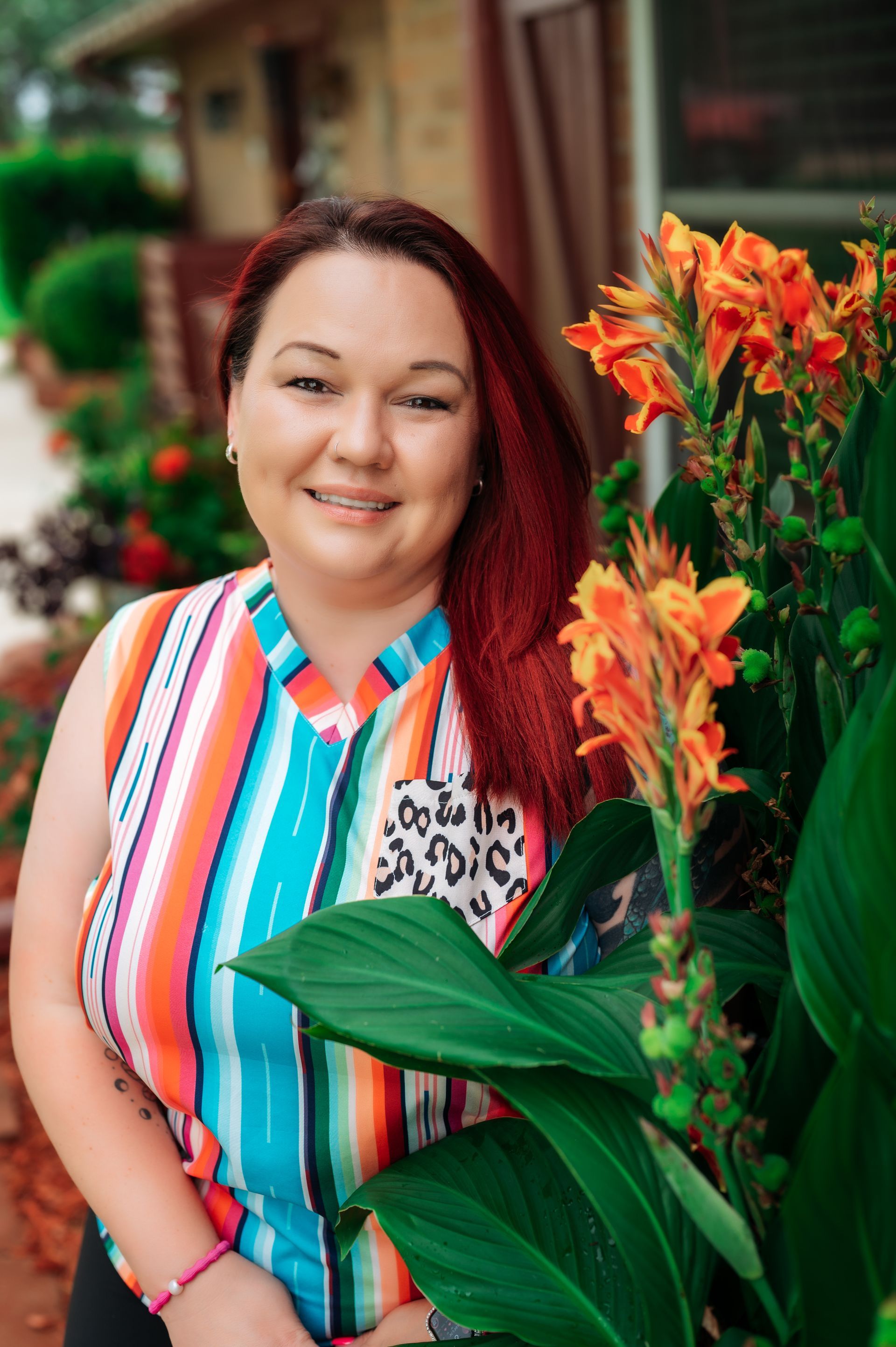 Woman with red hair smiling, wearing a colorful striped shirt, beside orange flowers.
