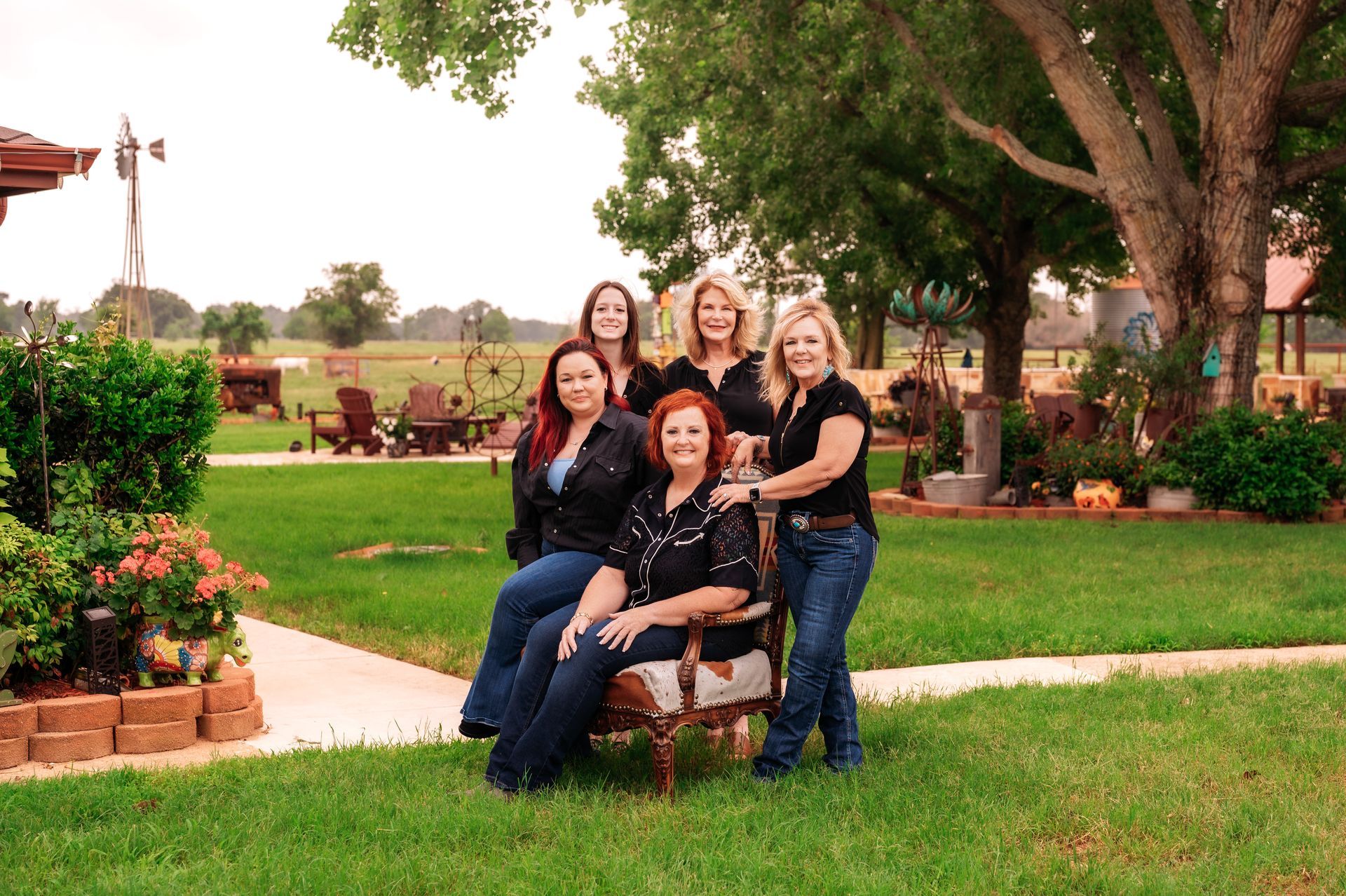 Six women in jeans and black shirts pose outdoors in a yard.
