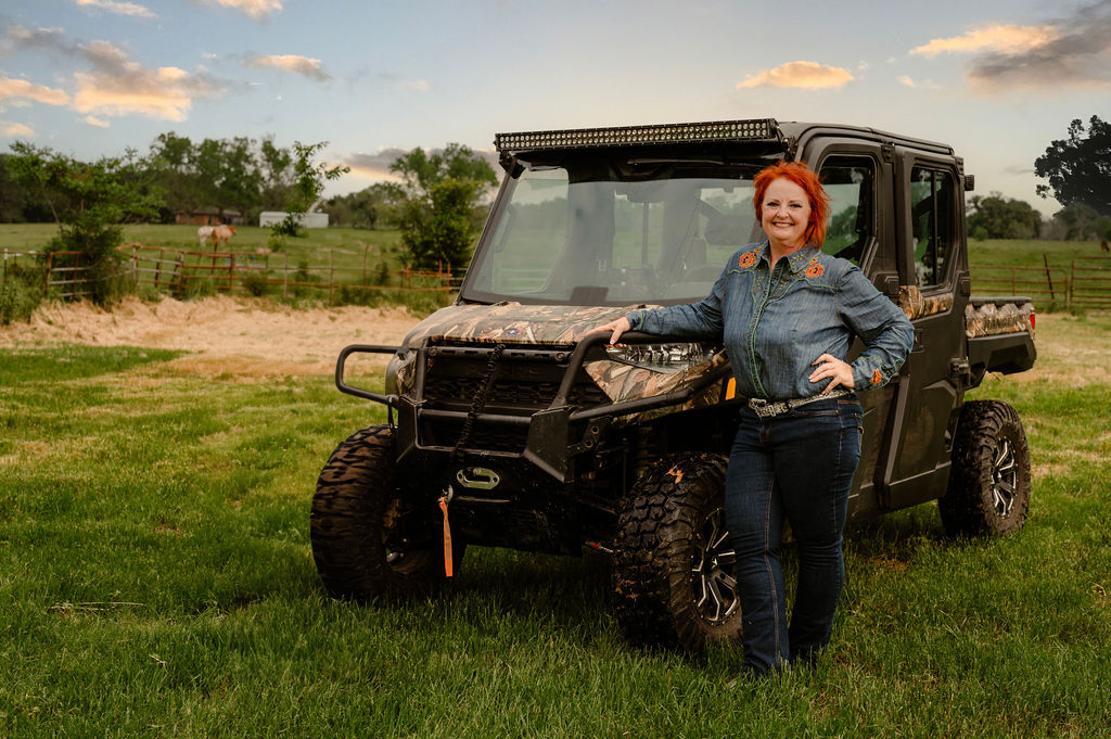 Woman in denim leans against a camouflage UTV in a grassy field.