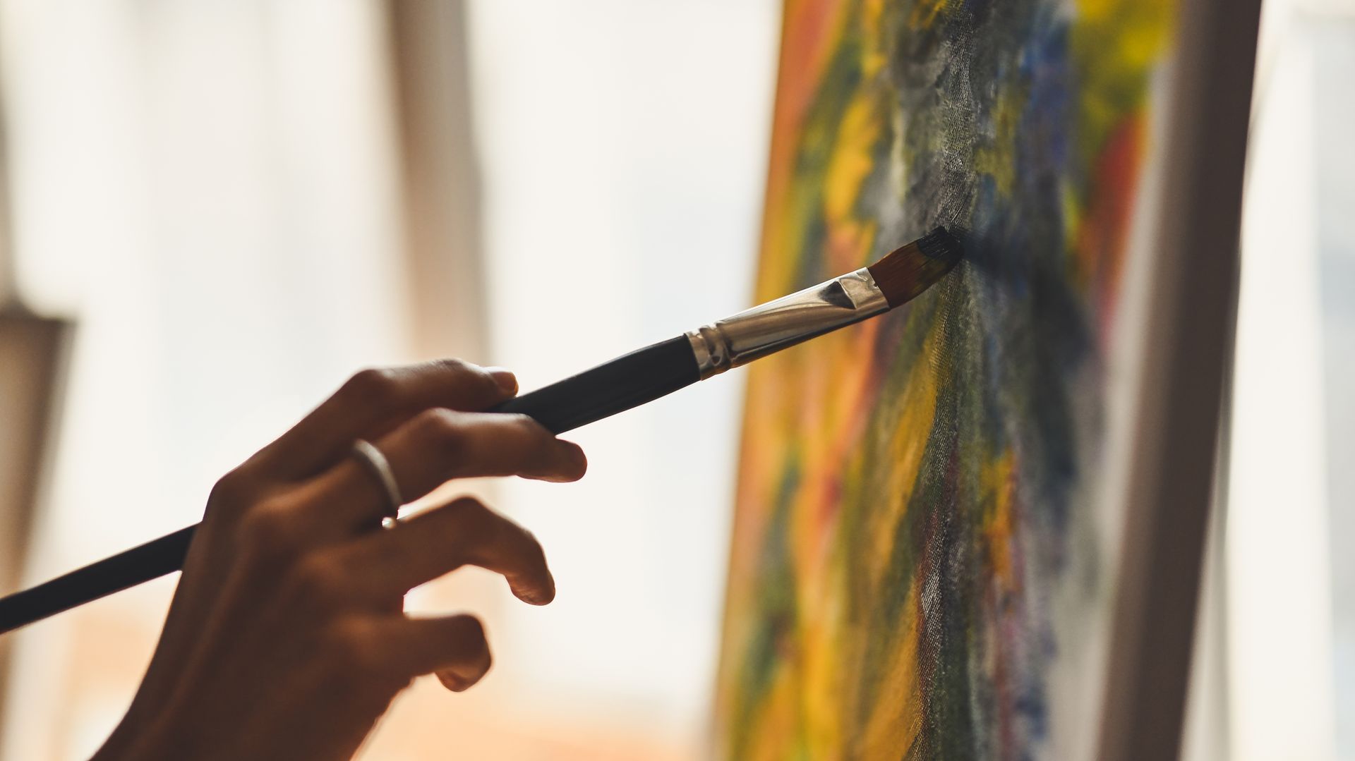 A close-up of a hand holding a paintbrush, applying dark blue paint to a colorful abstract canvas.
