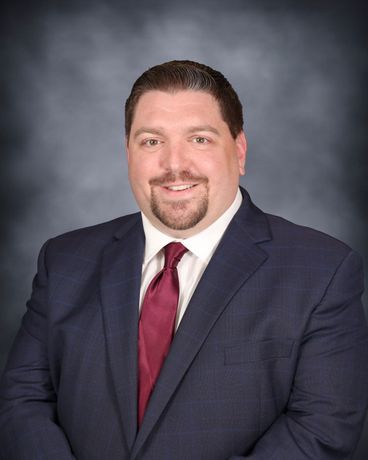 Professional headshot of a man in a navy suit, light blue shirt, and pink tie on a gray background