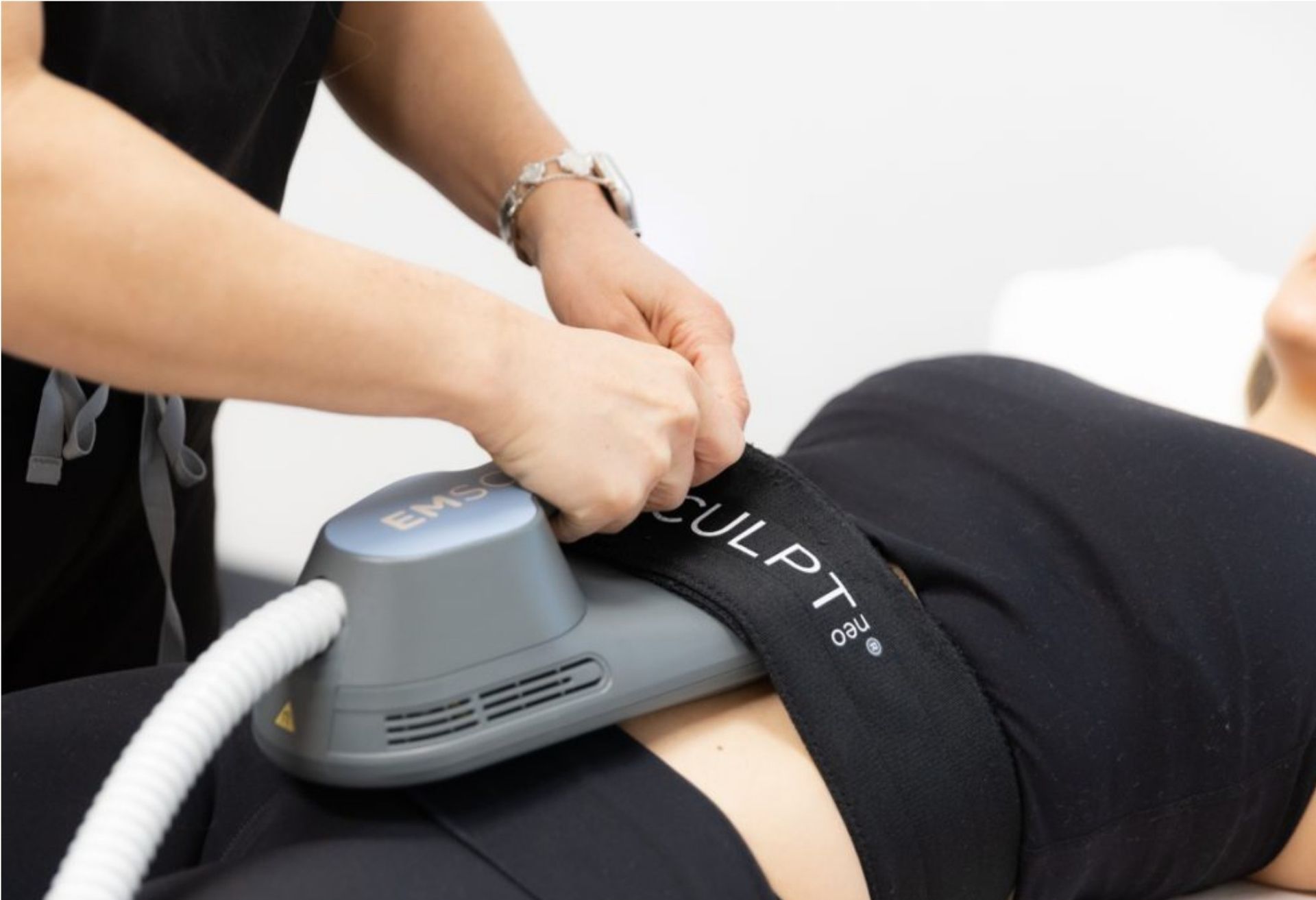 Person receiving EMSCULPT treatment on abdomen, in a medical office. Technician securing the device.