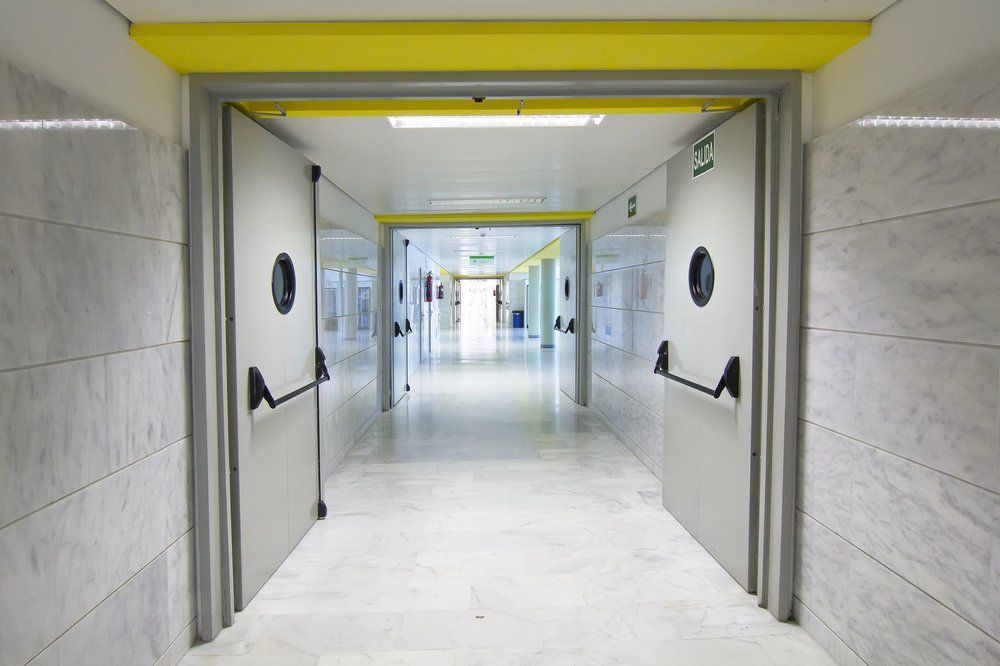 Couloir avec portes grises, fenêtres rondes et barres de poussée noires, éclairage vif au plafond.