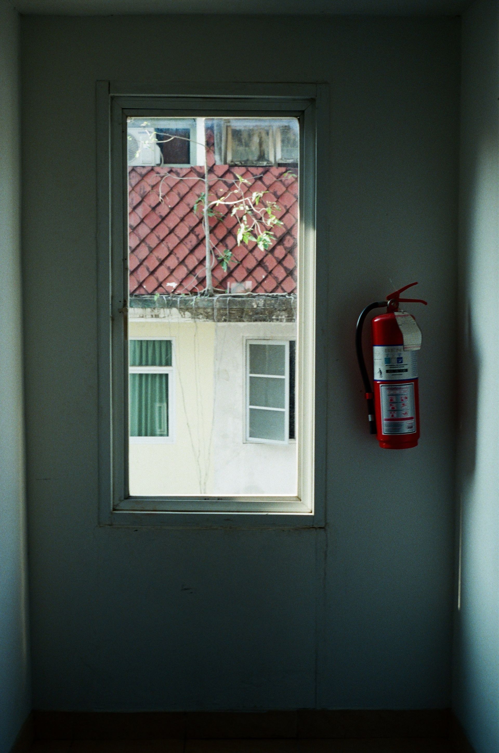 Vue par la fenêtre sur un toit de tuiles rouges, des bâtiments blancs et un extincteur fixé au mur.