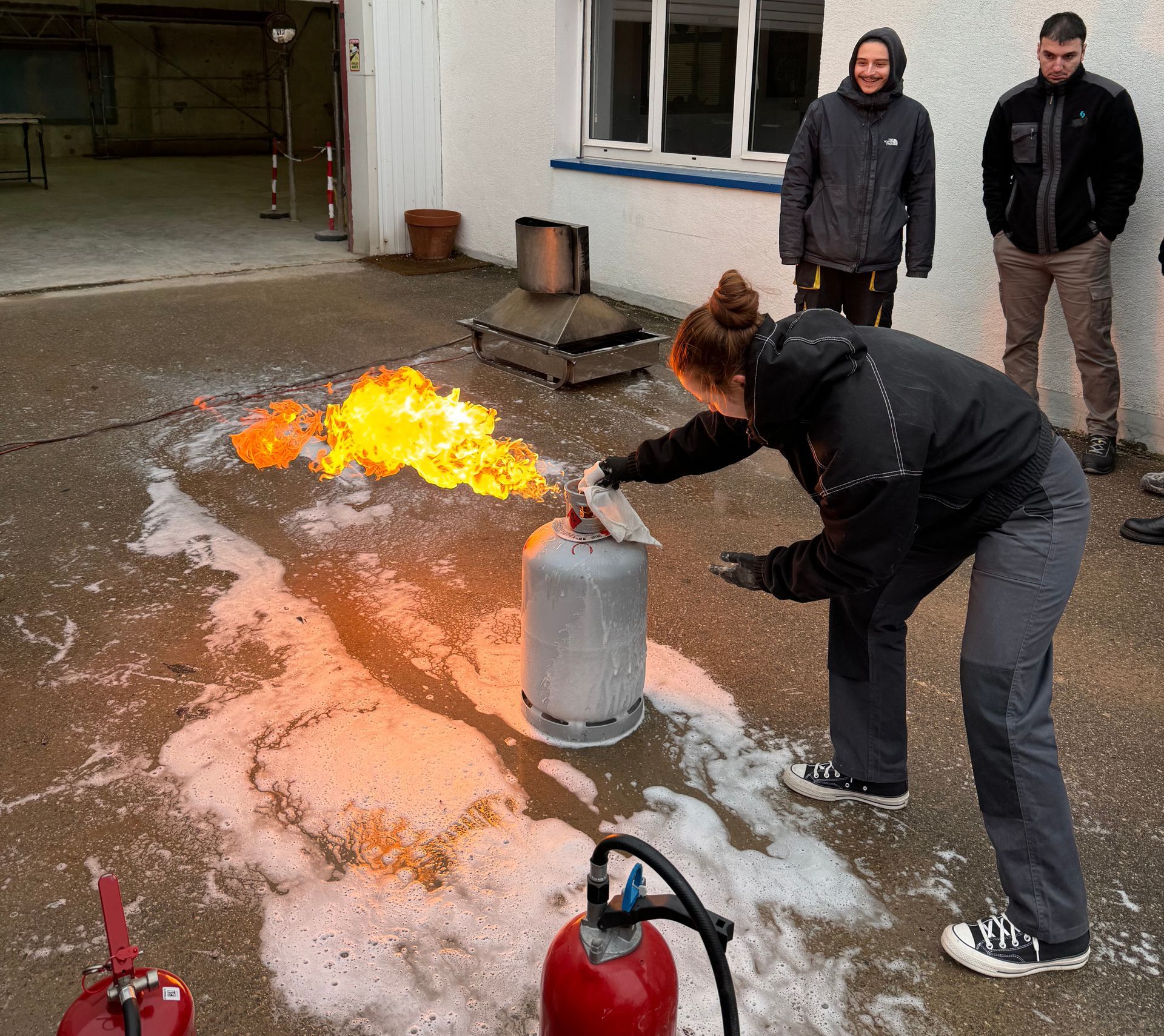 Une femme éteint un feu provenant d'une bonbonne de propane à l'aide d'un extincteur, sous le regard de deux hommes.