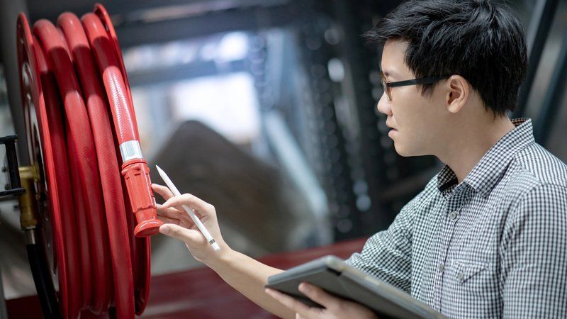 Un homme portant des lunettes inspecte un dévidoir de tuyau d'incendie rouge à l'aide d'une tablette, à l'intérieur.