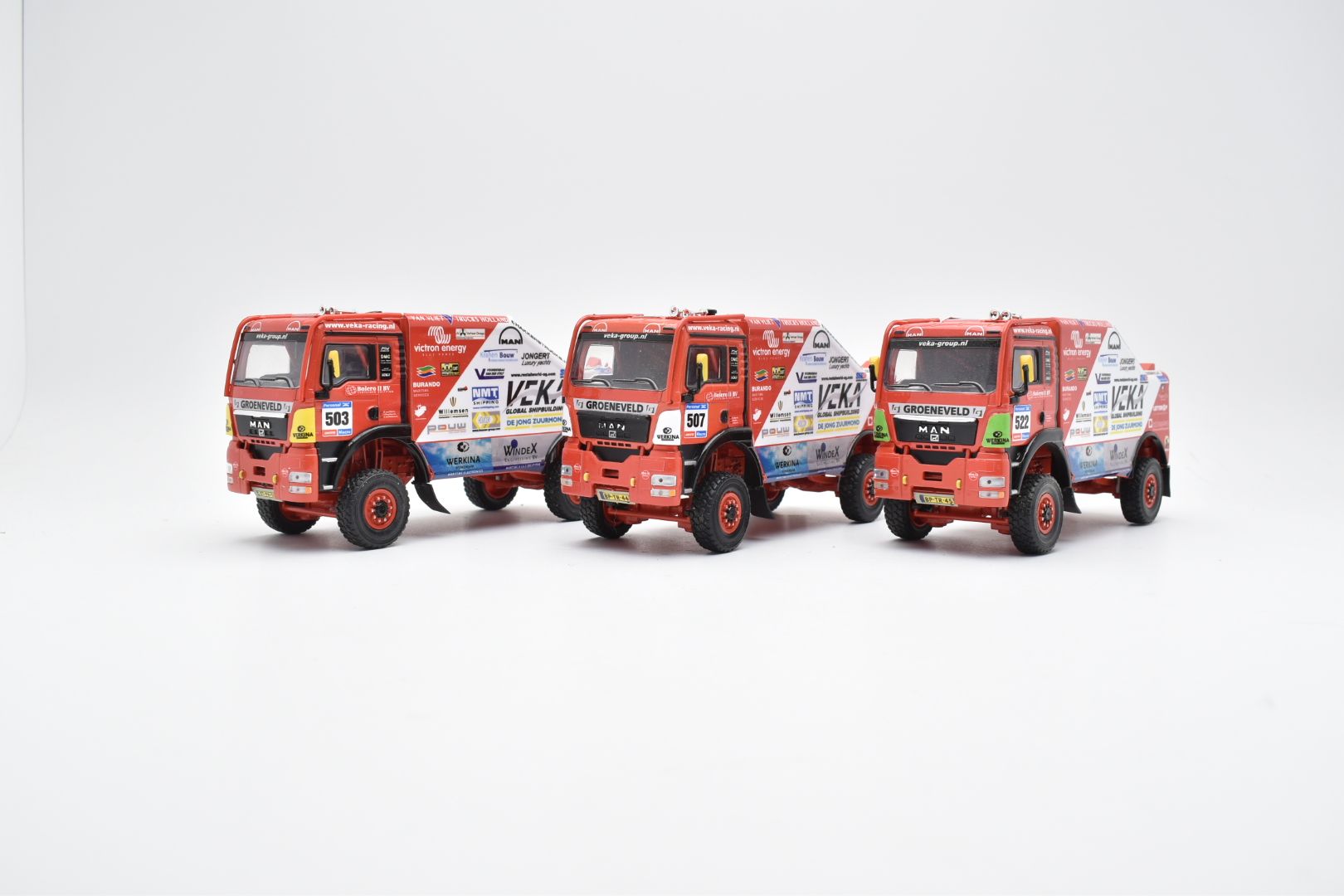 Drie rode rallytrucks op een witte achtergrond. Alle drie met sponsorlogo's en een vergelijkbaar ontwerp.