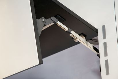 A metal kitchen cabinet hinge mechanism holding a white cupboard door partially open.