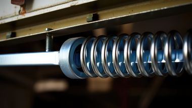 A close-up view of a metallic spring coiled around a blue rod, positioned beneath a metal beam.